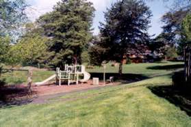 The path passes a tot lot next to a pond.