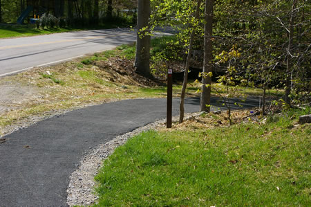 The concrete sidewalk ends and merges into an asphalt trail that goes into the woods.