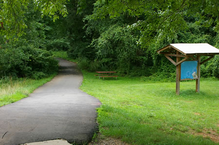 The concrete sidewalk leads into an asphalt trail that enters a wooded area.