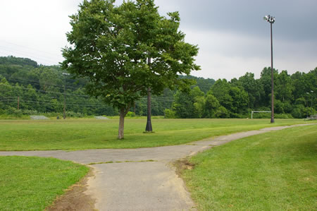 A trail intersects from the left at the end of a soccer field. Take the trail to the right to keep the soccer field on your left.