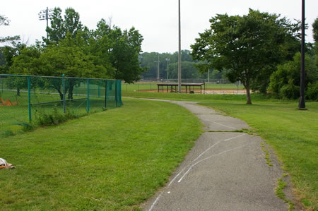 A trail intersects from the right at the end of a soccer field.  Continue on the present trail as it turns to the left.