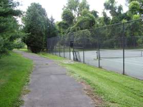 The trail passes a tennis court on the right and turns left to pass between the houses.