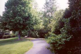 The trail emerges from between the homes with a grassy field on the left and homes on the right.